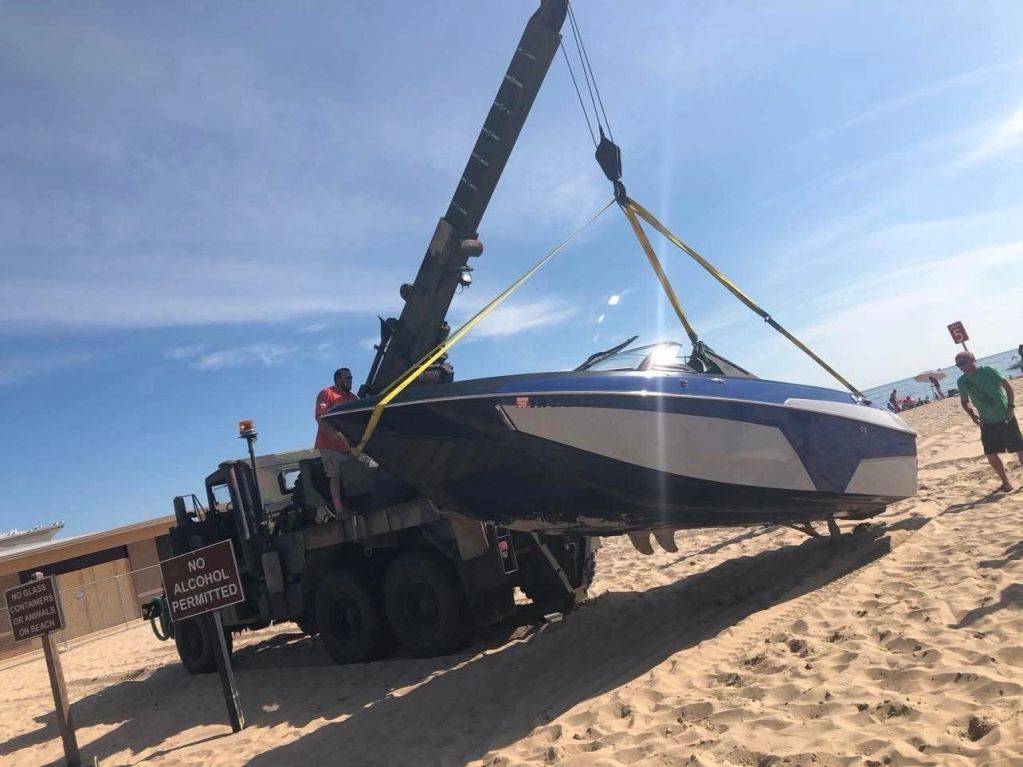 H2O Boat Tow's crane and crew hoisting a boat onto a transport truck at a beach location.