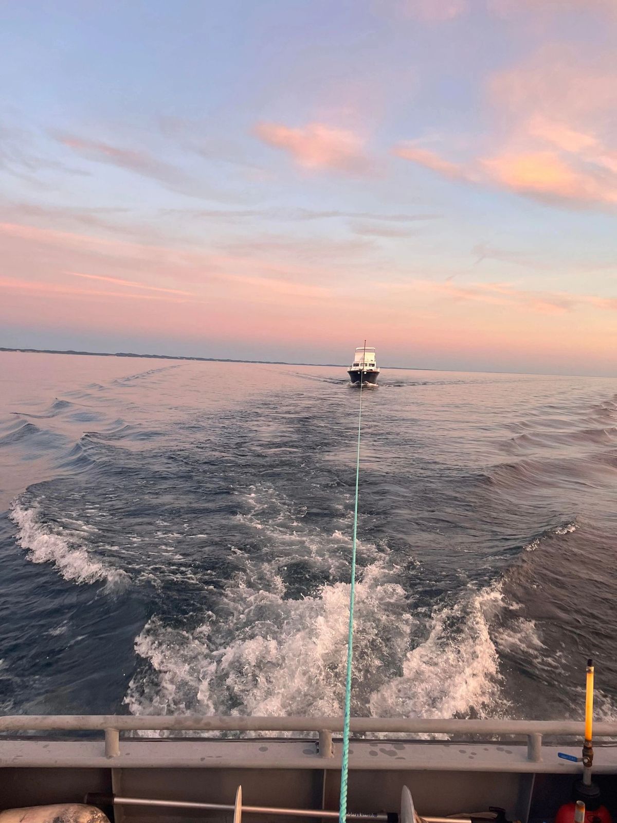 A boat towing another vessel across calm water at sunset, the rope line visible from the tow boat.