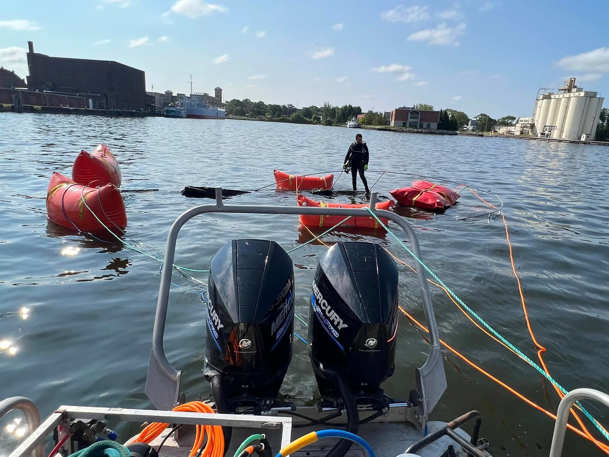 A diver working among lift bags set around a submerged boat, viewed from the towboat, with the waterfront in the background.