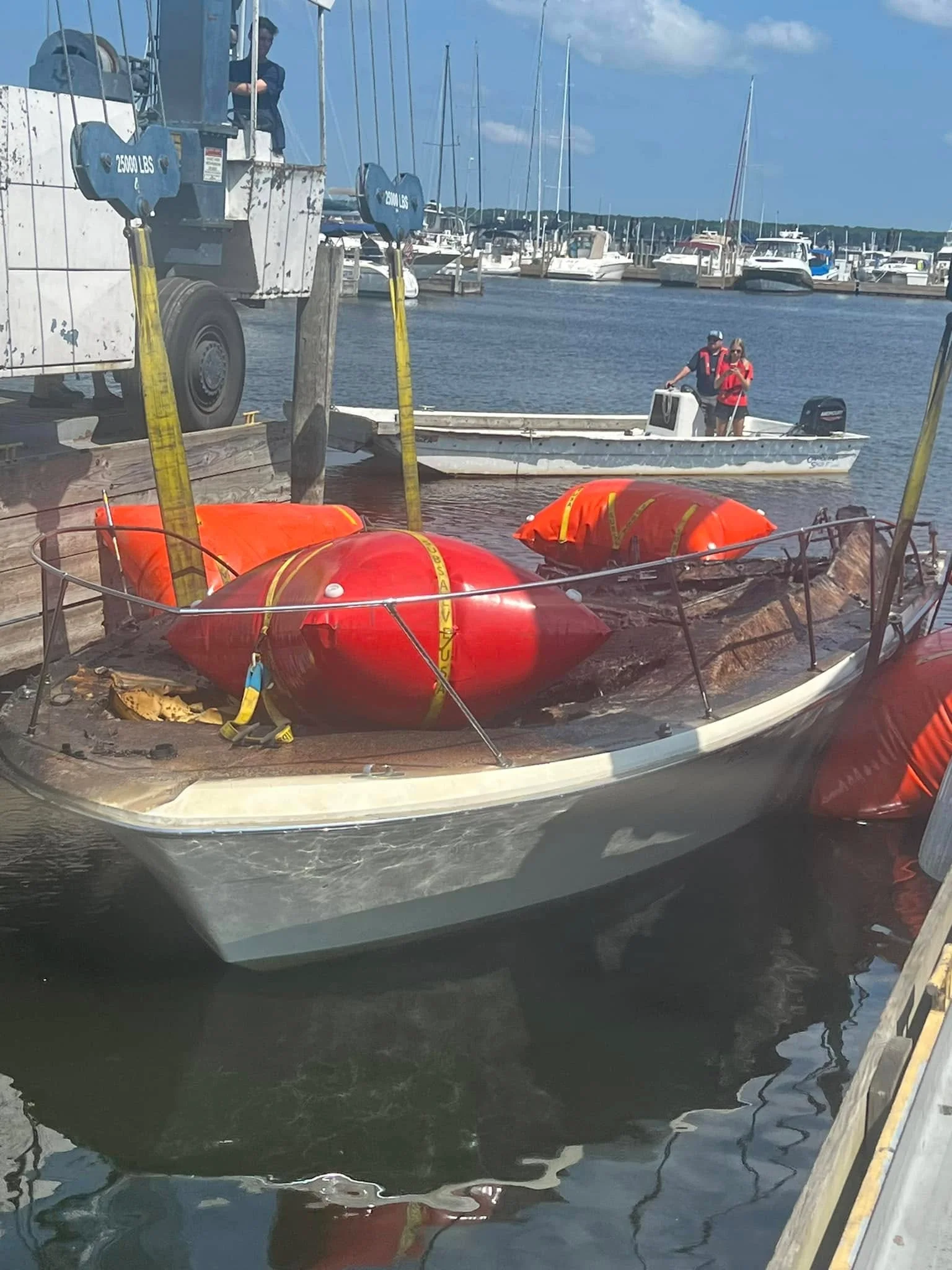 A crane hoisting a boat out of the water with lift bags still lashed to the deck, marina boats in the background.