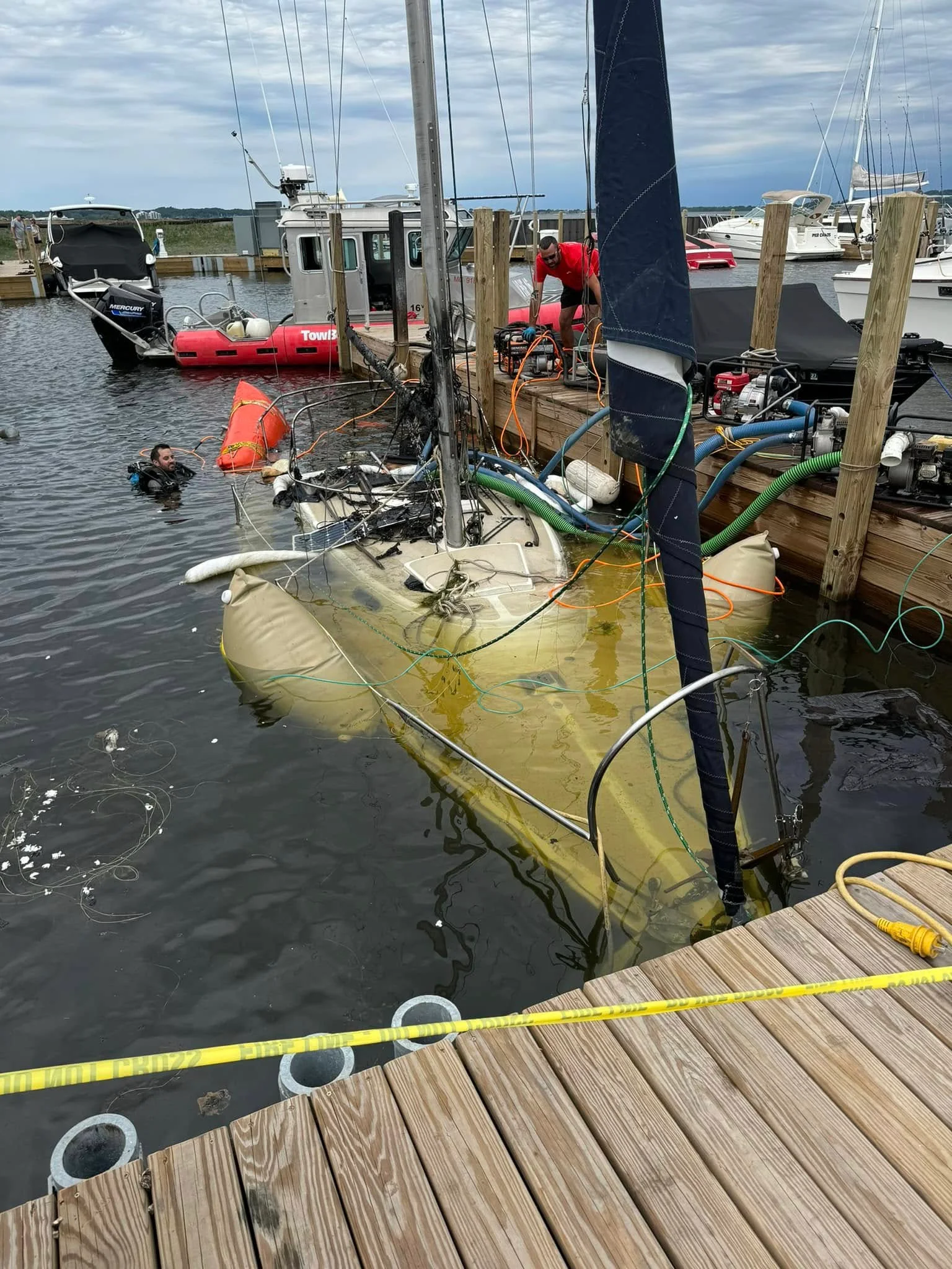 A diver working beside a partially submerged sailboat held up by lift bags, with pumps and hoses running on the dock.